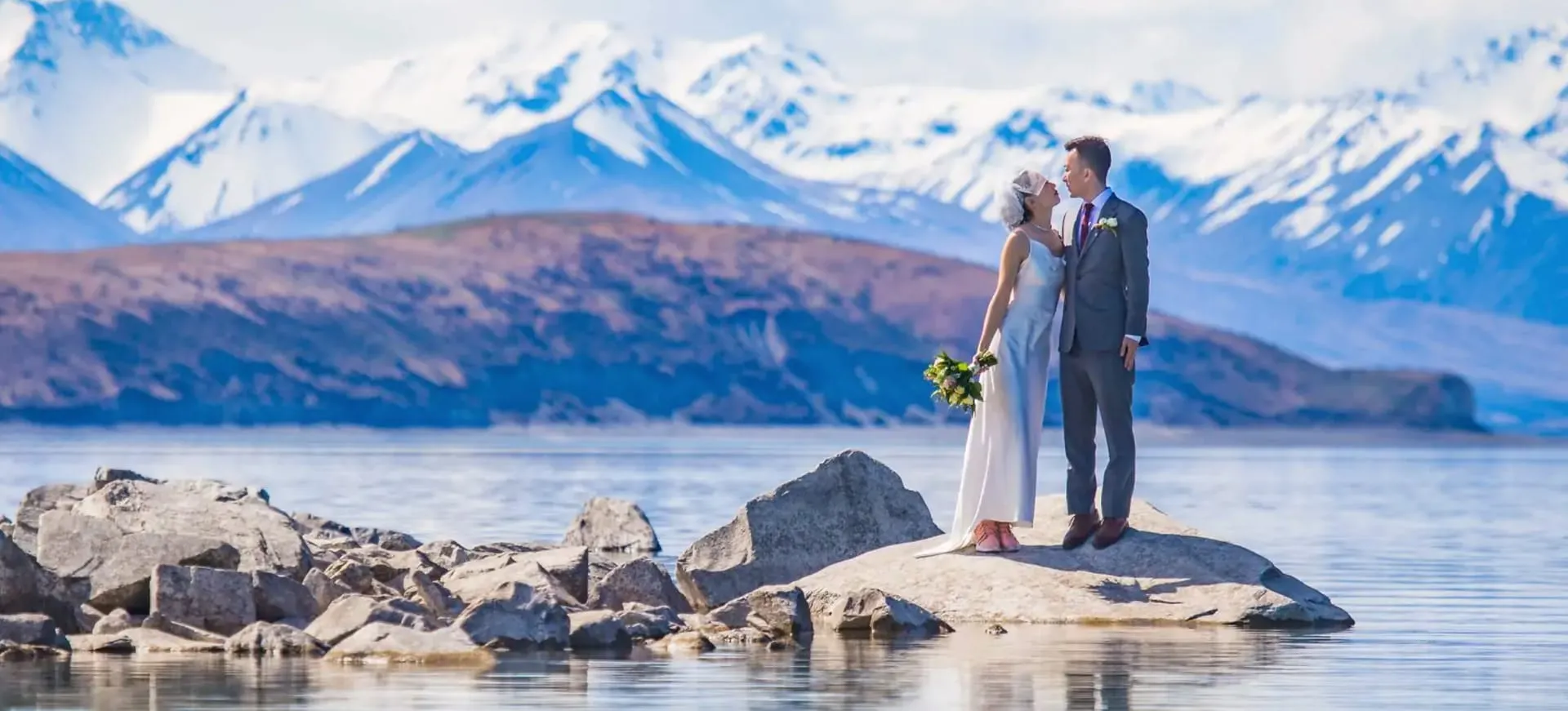 A couple on a rock in Lake Tekapo, framed by the snow-capped Southern Alps