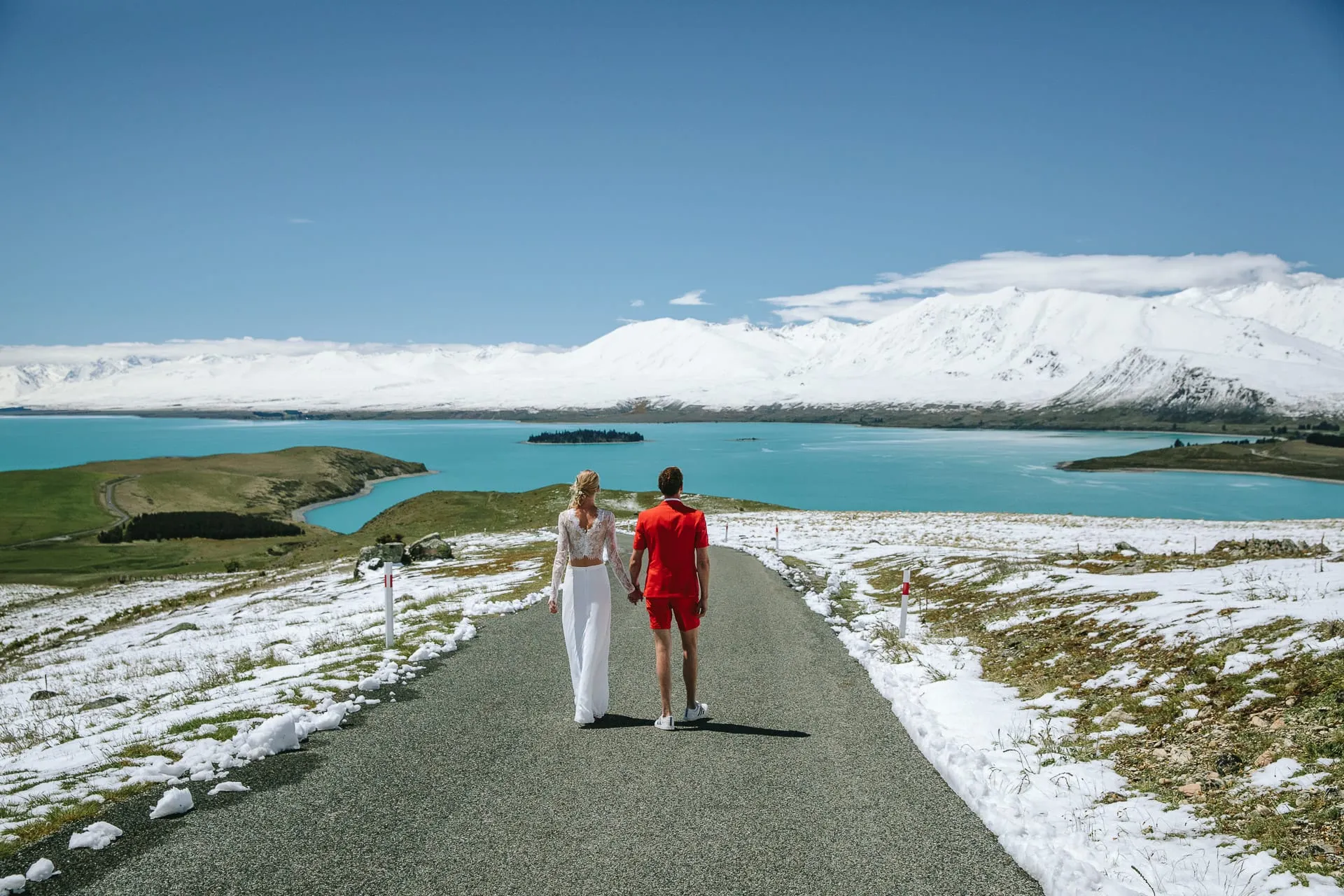 A couple walking hand in hand down a snow-lined road above turquoise Lake Tekapo, snow-capped mountains beyond