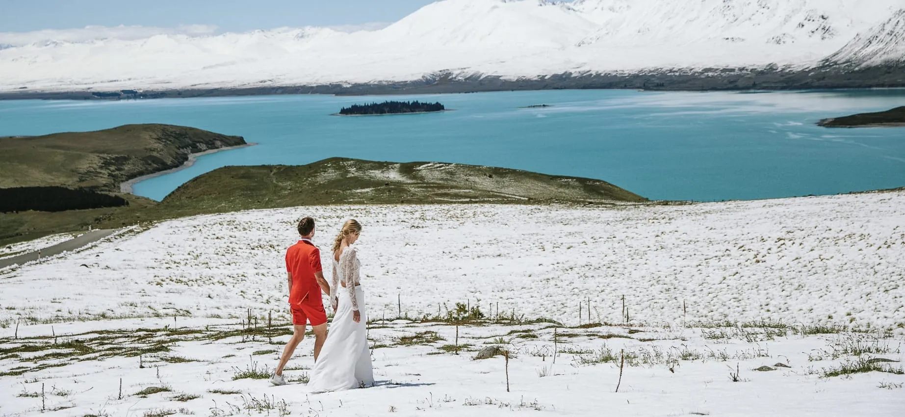 A couple walking across a snow-covered field above Lake Tekapo, with snow-capped mountains behind
