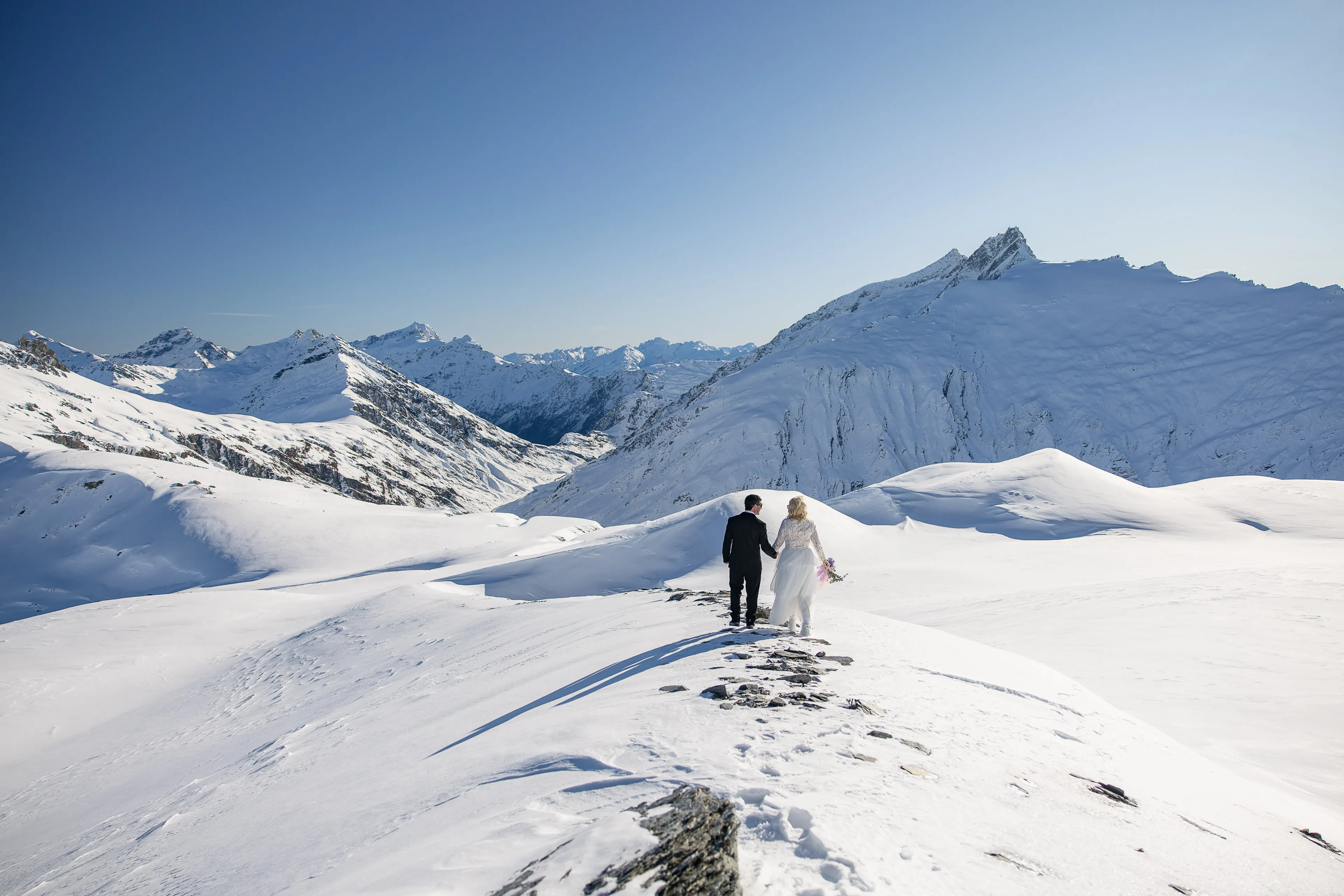A bride and groom walking across the snow on Tyndall Glacier, surrounded by the Southern Alps